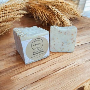 Könnte beinhalten: Ein Stapel handgemachter Honig- und Hafer-Bienenwachssoap mit einem Etikett mit der Aufschrift "Mersea Mudd Shop - Honey & Oat Beeswax Soap". Die Seife ist hellbeige und hat eine dezente Textur.