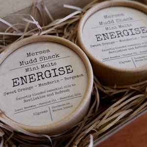 K&ouml;nnte beinhalten: Zwei kleine braune Pappbeh&auml;lter mit wei&szlig;en Etiketten. Die Etiketten lauten "Mersea Mudd Shack Mini Melts ENERGISE Sweet Orange - Mandarin - Bergamot Sorgf&auml;ltig ausgew&auml;hlte &auml;therische &Ouml;le zum Revitalisieren und Erfrischen Bienenwachs aus der Region - Kokosnuss&ouml;l - &Auml;therische &Ouml;le 50 Gramm Handgefertigt auf der Insel Mersea".