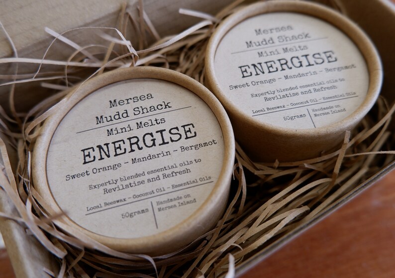K&ouml;nnte beinhalten: Zwei kleine braune Pappbeh&auml;lter mit wei&szlig;en Etiketten. Die Etiketten lauten "Mersea Mudd Shack Mini Melts ENERGISE Sweet Orange - Mandarin - Bergamot Sorgf&auml;ltig ausgew&auml;hlte &auml;therische &Ouml;le zum Revitalisieren und Erfrischen Bienenwachs aus der Region - Kokosnuss&ouml;l - &Auml;therische &Ouml;le 50 Gramm Handgefertigt auf der Insel Mersea".