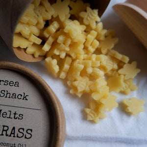 K&ouml;nnte beinhalten: Ein brauner Pappbeh&auml;lter mit dem Text "Mersea Judd Shack" und "Mini Melts" auf dem Etikett. Der Beh&auml;lter ist gef&uuml;llt mit kleinen, gelben Wachs-Schmelzen in Form von Sternen und Bienen. Die Wachsschmelzen fallen aus dem Beh&auml;lter auf eine wei&szlig;e Oberfl&auml;che.