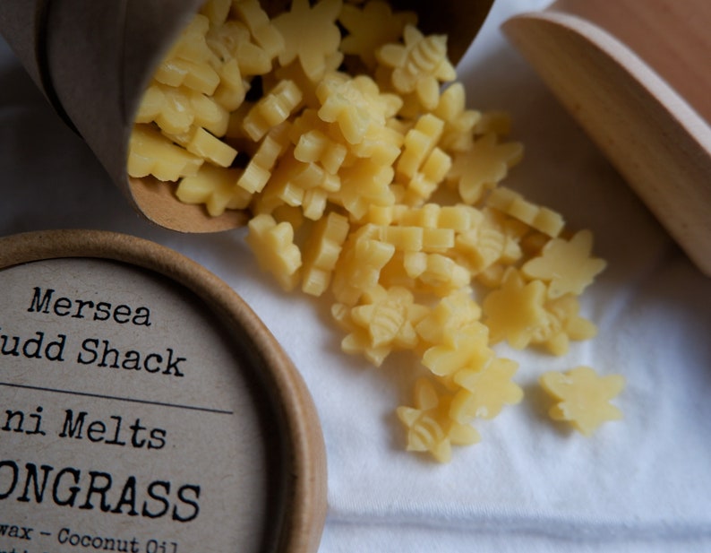 K&ouml;nnte beinhalten: Ein brauner Pappbeh&auml;lter mit dem Text "Mersea Judd Shack" und "Mini Melts" auf dem Etikett. Der Beh&auml;lter ist gef&uuml;llt mit kleinen, gelben Wachs-Schmelzen in Form von Sternen und Bienen. Die Wachsschmelzen fallen aus dem Beh&auml;lter auf eine wei&szlig;e Oberfl&auml;che.
