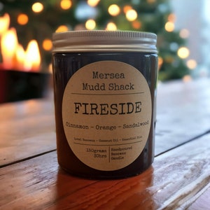 Könnte beinhalten: Ein Glas-Kerze mit silbernem Deckel und braunem Etikett. Auf dem Etikett steht "Mersea Mudd Shack FIRESIDE" mit Duftnoten von Zimt, Orange und Sandelholz. Die Kerze ist handgegossen und enthält 150 Gramm Bienenwachs.