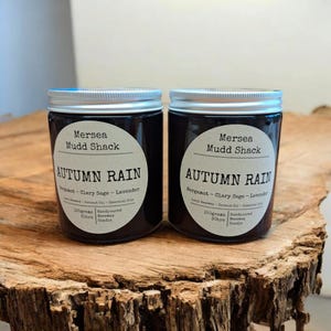 Könnte beinhalten: Zwei dunkle Gläser mit silbernen Deckeln, beschriftet mit "Mersea Mudd Shack AUTUMN RAIN" mit Duftdetails. Jede Kerze wiegt 150 Gramm und ist handgegossen. Die Gläser stehen auf einer rustikalen, strukturierten Holzoberfläche.