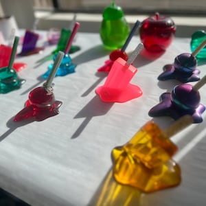 May include: A collection of colorful, melted candy-like objects on a white surface. The objects are shaped like lollipops, apples, popsicles, and other abstract shapes. Some of the objects have sticks or skewers inserted into them.