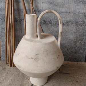 May include: A white ceramic vase with a unique, rounded shape and a single handle. The vase has a textured surface and a slightly distressed look.