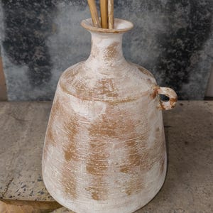 Puede incluir: Un jarrón de cerámica blanca con un esmalte marrón y un asa. El jarrón tiene una base ancha y un cuello estrecho.