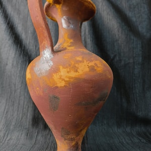 May include: A tall, terracotta-colored vase with a curved handle and a wide, flared rim. The vase has a bulbous body and a narrow base. The surface has a weathered, aged appearance with shades of brown, orange, and yellow, suggesting an antique or rustic aesthetic.