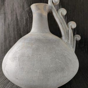 Peut inclure: Une cruche en céramique grise avec un corps bulbeux et un col fin. La poignée présente un motif décoratif en spirale. La cruche a une surface texturée et un bord légèrement plus foncé.