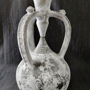 May include: A weathered, off-white ceramic amphora with two handles and a flared top. The vessel has a bulbous base and a textured surface with dark gray and black markings, suggesting age and use. The design includes a central spout and two small decorative knobs.