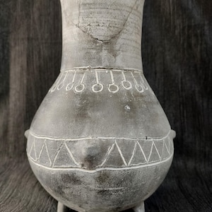 Puede incluir: Un jarrón de cerámica gris con un cuerpo ancho y redondeado y un cuello alto y acampanado. El jarrón tiene tres pequeños pies y dos pequeñas asas. Patrones geométricos blancos y una serie de círculos decoran el cuerpo. El jarrón tiene un aspecto envejecido.