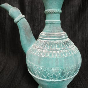 May include: A turquoise ceramic pitcher with a spout and handle. The pitcher features detailed white embossed patterns around the body and neck, with a rope-like design near the top. The base is round and the overall shape is bulbous.