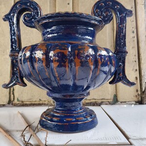 Puede incluir: Un jarrón de cerámica azul con dos asas ornamentadas. El jarrón tiene un cuerpo redondeado y una base acampanada. Las asas están curvadas y tienen un diseño decorativo. El jarrón tiene un acabado desgastado con un toque de cobre.
