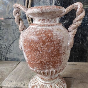 Puede incluir: Un jarrón de color terracota con una superficie texturizada y detalles en blanco. El jarrón presenta dos asas trenzadas en forma de cuerda y una base ornamentada y detallada. El diseño general tiene una apariencia envejecida y antigua.