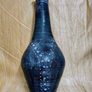 May include: A tall, dark blue vase with a textured surface and a speckled pattern of white dots. The vase has a slender neck and a rounded body, suitable for displaying flowers or as a decorative piece.