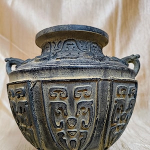 以下が含まれることがあります： 複雑なレリーフ模様と2つの小さな取っ手が付いた、濃い灰色のアンティークスタイルの花瓶。花瓶は幅広で丸みを帯びたボディと、わずかにフレアしたリムを持っています。表面は風化した外観で、古さと歴史を示唆しています。