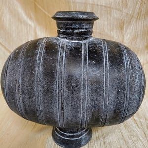 Puede incluir: Un jarrón negro texturizado con un cuerpo redondeado y un cuello estrecho. Líneas verticales recorren el cuerpo del jarrón, y se apoya sobre una pequeña base circular. El jarrón tiene un acabado mate y parece estar hecho de piedra o un material similar.