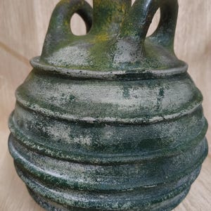 Puede incluir: Una jarra de cerámica verde oscuro y desgastada, con cuerpo redondo y dos asas arqueadas. La jarra tiene una textura acanalada alrededor del cuerpo y una pequeña abertura cilíndrica en la parte superior. Se ven manchas amarillas y verdes.