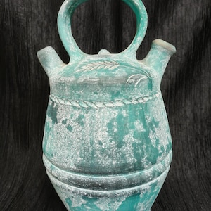 May include: A weathered, turquoise-colored ceramic water jug with a large handle and two spouts. The jug features a decorative rope-like band around the middle and embossed wheat-like designs. The jug has a distressed, aged appearance.