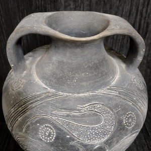 May include: A gray ceramic vase with two handles and a wide, fluted rim. The vase features etched designs of swirling lines and stylized floral or aquatic motifs. The overall aesthetic is rustic and antique-inspired.