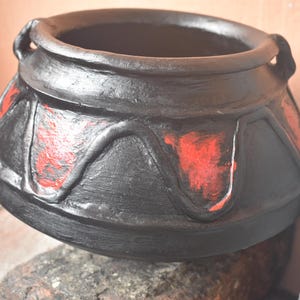 Puede incluir: Olla de barro negro con detalles rojos. La olla tiene una boca ancha y un cuerpo redondeado. Está decorada con un patrón ondulado alrededor del borde.