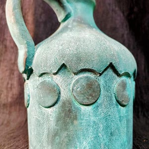 May include: A teal-colored ceramic jug with a handle and a decorative pattern of circles and zigzags. The jug has a textured surface and a weathered appearance, suggesting an antique or vintage style.