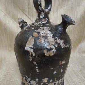 Puede incluir: Una jarra de cerámica oscura de estilo antiguo, con asa y dos picos. La jarra es principalmente negra con zonas de pintura desgastada blanquecina, lo que le da un aspecto rústico. El asa es un gran lazo, y los picos son pequeños e inclinados.