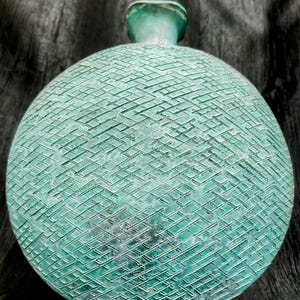 May include: A round, turquoise decorative vase with a textured, cross-hatched pattern. The vase has a small, cylindrical neck and a flat base. The design is reminiscent of a vintage or antique piece.