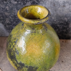 Könnte beinhalten: Eine handgefertigte, kugelförmige Vase mit einer breiten, kreisförmigen Öffnung. Die Vase hat eine strukturierte Oberfläche mit einer Mischung aus grünen, gelben und schwarzen Farbtönen. Der Rand ist in einem dunkleren Gelbton gehalten, und das Gesamtdesign ist rustikal und einzigartig.