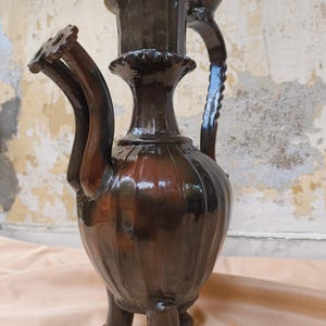 Puede incluir: Una jarra de cerámica marrón oscuro con un cuerpo bulboso y tres patas. La jarra tiene un cuello alto y cilíndrico y un asa curva. Dos picos con un diseño en forma de engranaje se extienden desde el lateral. La superficie tiene un acabado brillante.