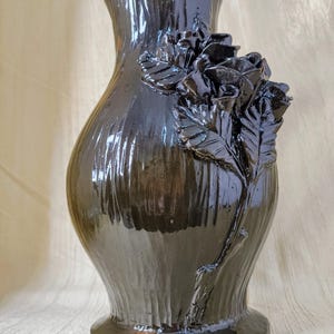 May include: A glossy, dark brown ceramic vase with a sculpted rose design. The vase has a flared top and a wide base, with vertical ridges adding texture. The roses are clustered on the side, adding a decorative element to the vessel.