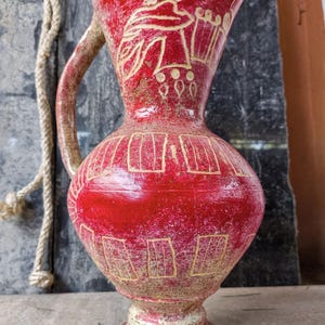 May include: A red ceramic jug with a handle and a wide base. The jug features a decorative design in a lighter colour, with a figure and geometric patterns. A rope is visible in the background.