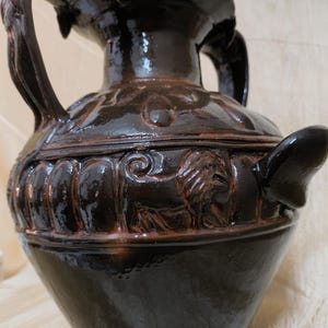 May include: A dark brown ceramic vase with a glossy finish. The vase features two handles and a spout. The body of the vase is decorated with relief carvings of lions and floral patterns. The vase is approximately 12 inches tall.