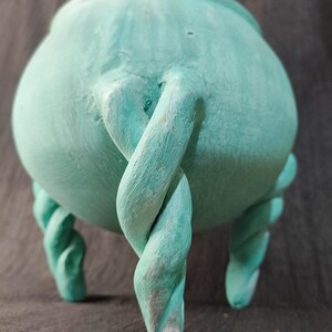 May include: A teal-colored ceramic cauldron with a rounded body and twisted rope-like legs. The cauldron has a textured surface and a twisted handle. The overall design is whimsical and decorative.