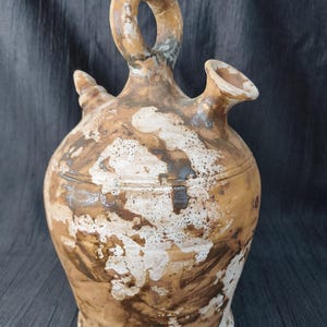 Puede incluir: Una jarra de agua rústica de cerámica con un diseño único. La jarra presenta un asa de bucle, un pico y una pequeña protuberancia decorativa. El exterior es principalmente marrón con acentos blancos y marrón oscuro, lo que le da una apariencia desgastada. La jarra mide aproximadamente 25 cm de altura.