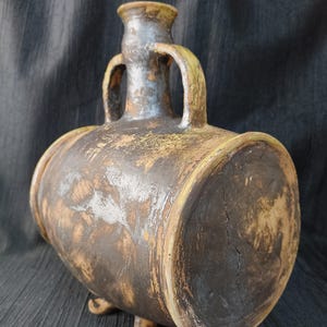 May include: A handcrafted, rustic ceramic vessel with a barrel-like shape, featuring two arched handles and three small feet. The pottery has a distressed, aged appearance with shades of brown, tan, and gray, suggesting an antique or vintage aesthetic.