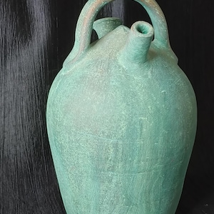 Puede incluir: Una jarra de cerámica verde desgastada, con asa y dos picos. La jarra tiene un cuerpo redondeado y una superficie texturizada, lo que sugiere una apariencia envejecida o rústica. El asa es de un tono marrón claro.