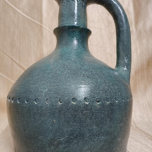 以下が含まれることがあります： 丸みを帯びたボディとハンドルが付いた、ターコイズ色のセラミックピッチャー。ピッチャーは、表面にテクスチャ加工が施されており、首の周りには小さな穴がいくつかあります。ハンドルは湾曲しており、ピッチャーの上部に取り付けられています。