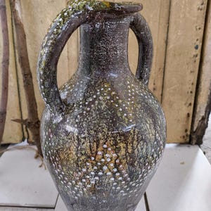 Puede incluir: Un jarrón de cerámica marrón con dos asas y un diseño moteado blanco. El jarrón tiene un fondo redondeado y un cuello estrecho.