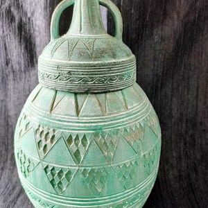 May include: A teal-colored, decorative ceramic vase with a lid and two handles. The vase features intricate geometric patterns and etched designs around the body and lid. The vase has a weathered, antique appearance.