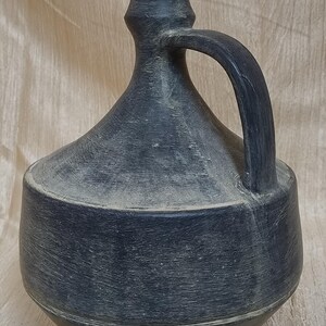 Puede incluir: Una jarra de cerámica azul oscuro con asa. La jarra tiene un cuerpo redondeado, un cuello estrecho y un borde acampanado. El asa es arqueada y está unida al cuello y al cuerpo. La superficie tiene un aspecto texturizado.