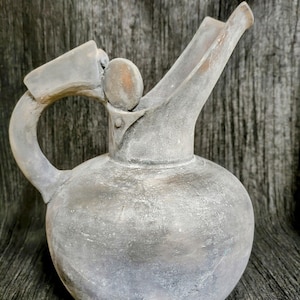 Puede incluir: Una jarra de cerámica gris con cuerpo redondeado, asa curva y pico. La jarra tiene una tapa con un pequeño pomo y una superficie texturizada. El diseño general es rústico y vintage.