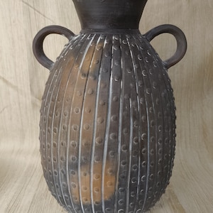 May include: A dark gray ceramic vase with two handles and a textured surface. The vase has a bulbous body with vertical lines and small, evenly spaced dots. The top is flared and the handles are curved.