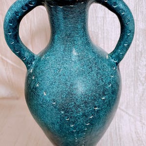 Puede incluir: Un jarrón de cerámica de color verde azulado con dos asas arqueadas. El jarrón tiene una superficie texturizada con pequeñas hendiduras espaciadas uniformemente. Las asas están adornadas con detalles circulares. El jarrón tiene una forma de ánfora clásica.