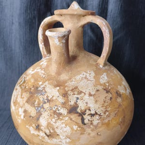 Puede incluir: Una jarra de cerámica de color marrón claro, de estilo antiguo, con cuerpo redondeado, dos asas arqueadas y un pico. La jarra tiene tapa y está decorada con patrones moteados marrones y blancos, que sugieren desgaste.