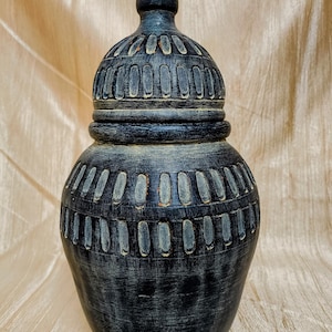 Può includere: Un vaso decorativo grigio scuro con coperchio. Il vaso ha un corpo arrotondato e una superficie strutturata con dettagli scolpiti verticali e orizzontali. Il coperchio è sormontato da un piccolo pinnacolo appuntito. Il vaso è probabilmente in ceramica o materiale simile.