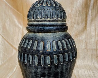 Op de afbeelding: Een donkergrijze decoratieve pot met een deksel. De pot heeft een afgeronde body en een textuur met verticale en horizontale gesneden details. Het deksel is bekroond met een kleine, puntige finial. De pot is waarschijnlijk gemaakt van keramiek of een vergelijkbaar materiaal.