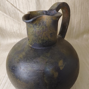 May include: A dark, round ceramic pitcher with a handle and a flared spout. The pitcher has a mottled finish in shades of black, green, and brown. The handle is arched and attached to the top of the pitcher.