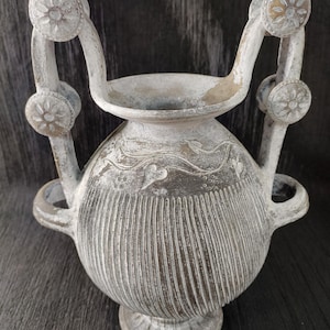 Op de afbeelding: Een verweerde, grijs getinte decoratieve urn met een ronde body en verticale ribbels. Het heeft twee gebogen handvatten met bloemmotieven en een voetstuk. Het ontwerp van de urn suggereert een antieke of klassieke stijl.