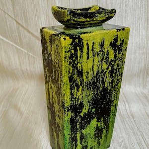 May include: A tall, rectangular vase with a textured, distressed finish in shades of green and black. The vase has a small, boat-shaped dish on top, also in green and black. The overall design is modern and artistic.