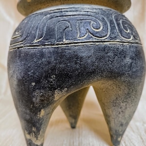 Può includere: Un bruciatore di incenso in ceramica grigio scuro a tre gambe. Il vaso ha un corpo arrotondato con una fascia decorativa di motivi scolpiti vicino alla parte superiore. Il bordo è di una tonalità di grigio più chiara. Il bruciatore ha un aspetto invecchiato.